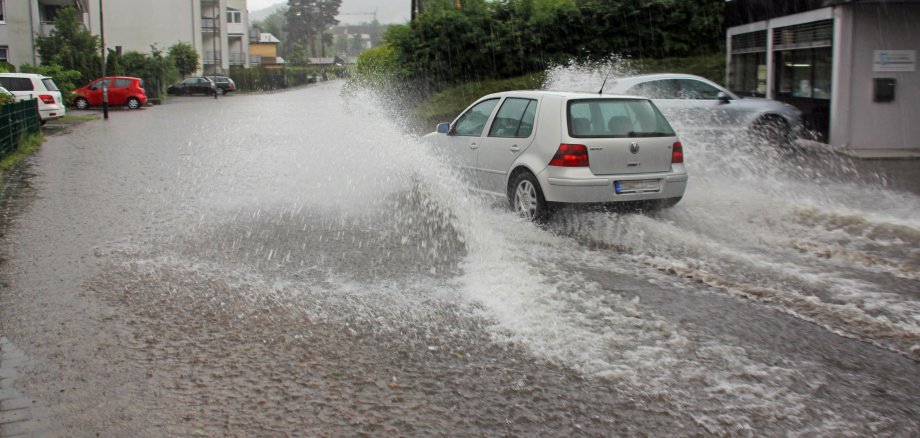 Ein Auto fährt im Starkregen durch eine überflutete Straße eines Wohngebietes